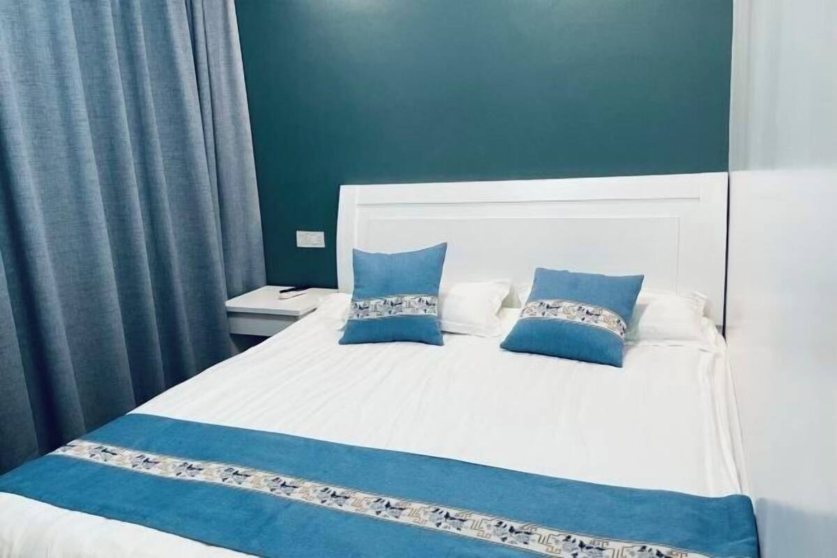 Ein ordentlich bezogenes Bett mit weißer Bettwäsche und blauen Akzenten, ein weißer Kopfteil und ein blauer Vorhang.