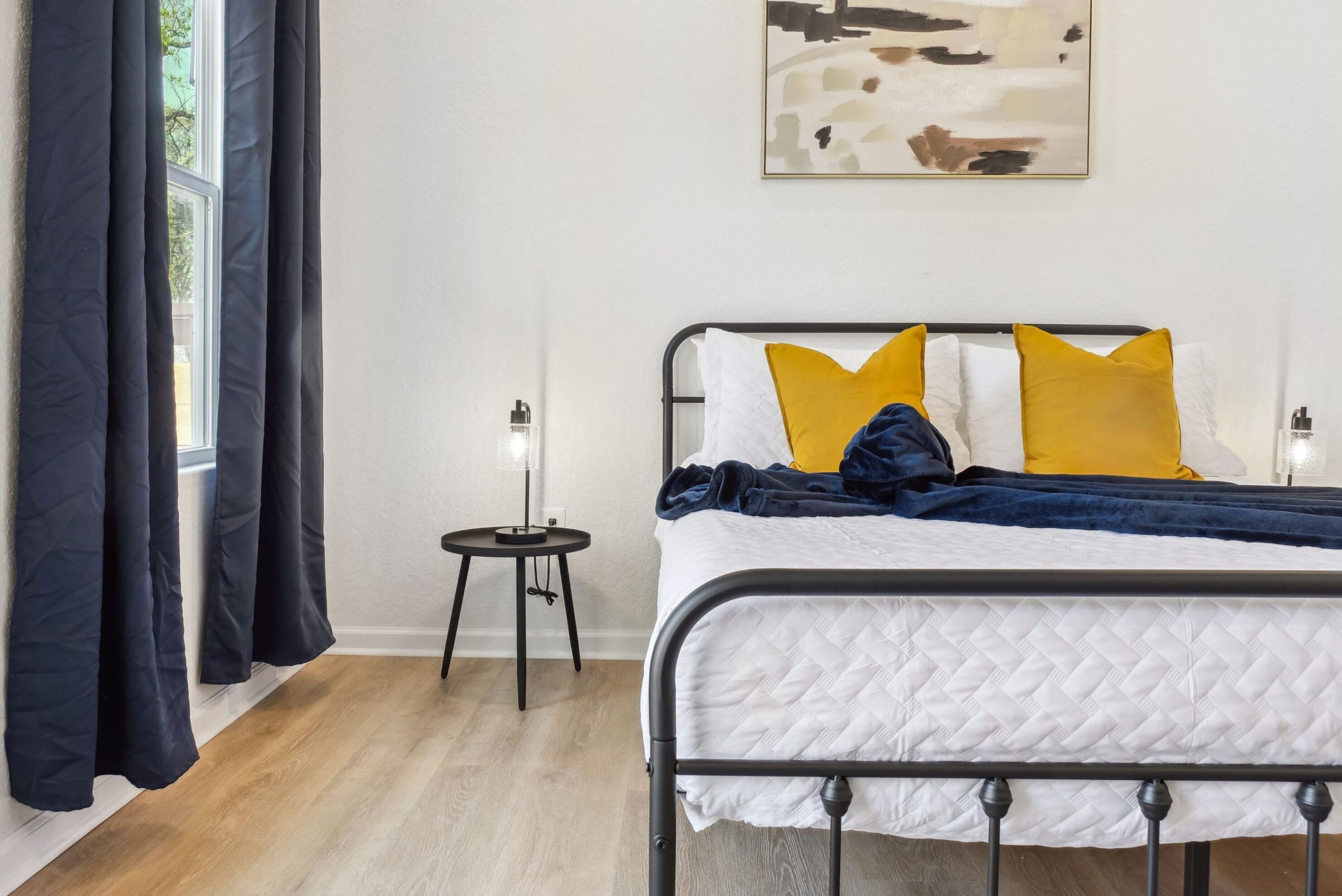 A neatly made bed with a white quilted bedspread, two yellow pillows, and a blue blanket. There is a small bedside table with a lamp, and a window with dark curtains.