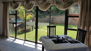 A dining area with a table set for two, large windows with curtains, and a view of a garden and pool. - El Shadai Guest House  Hartbeespoort (Hartbeespoort)