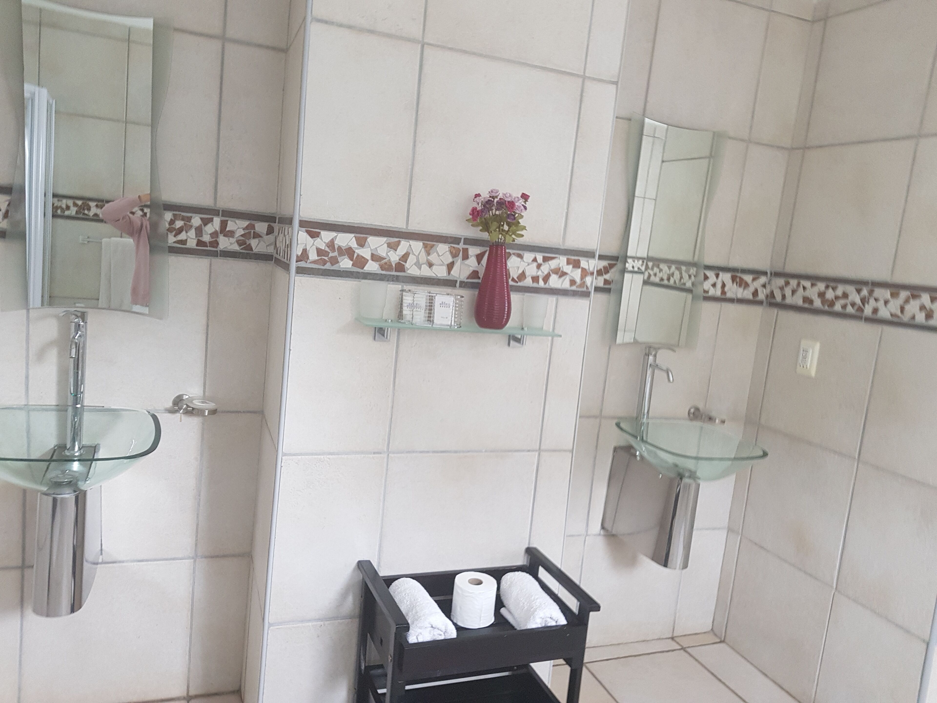 Casa de banho com duas pias de vidro, um espelho, um porta-toalhas, uma prateleira com artigos de higiene e um carrinho preto com toalhas.