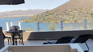 A wooden hot tub on a terrace with a table and chairs, overlooking a hillside with houses. - El Shadai Guest House  Hartbeespoort (Hartbeespoort)