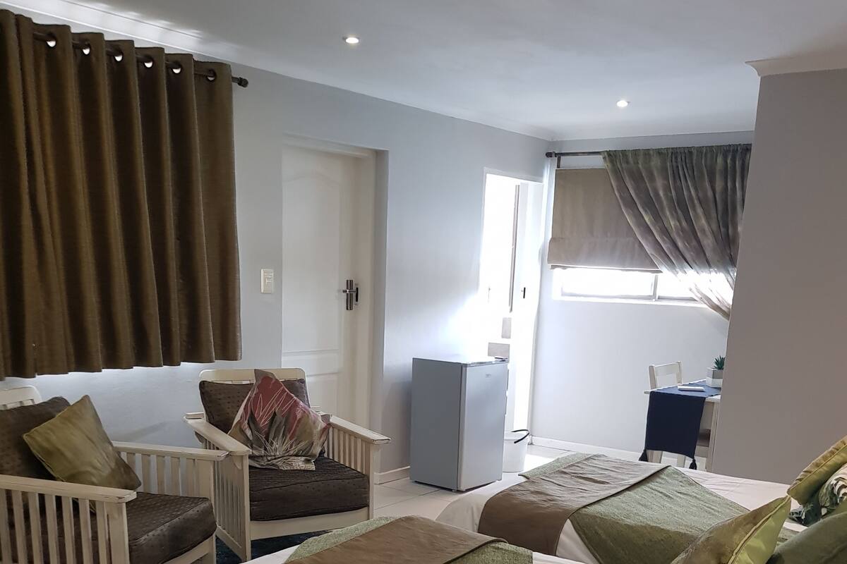 Quarto de hotel com duas camas, uma pequena escrivaninha, uma cadeira, um frigorífico e uma janela com cortinas.