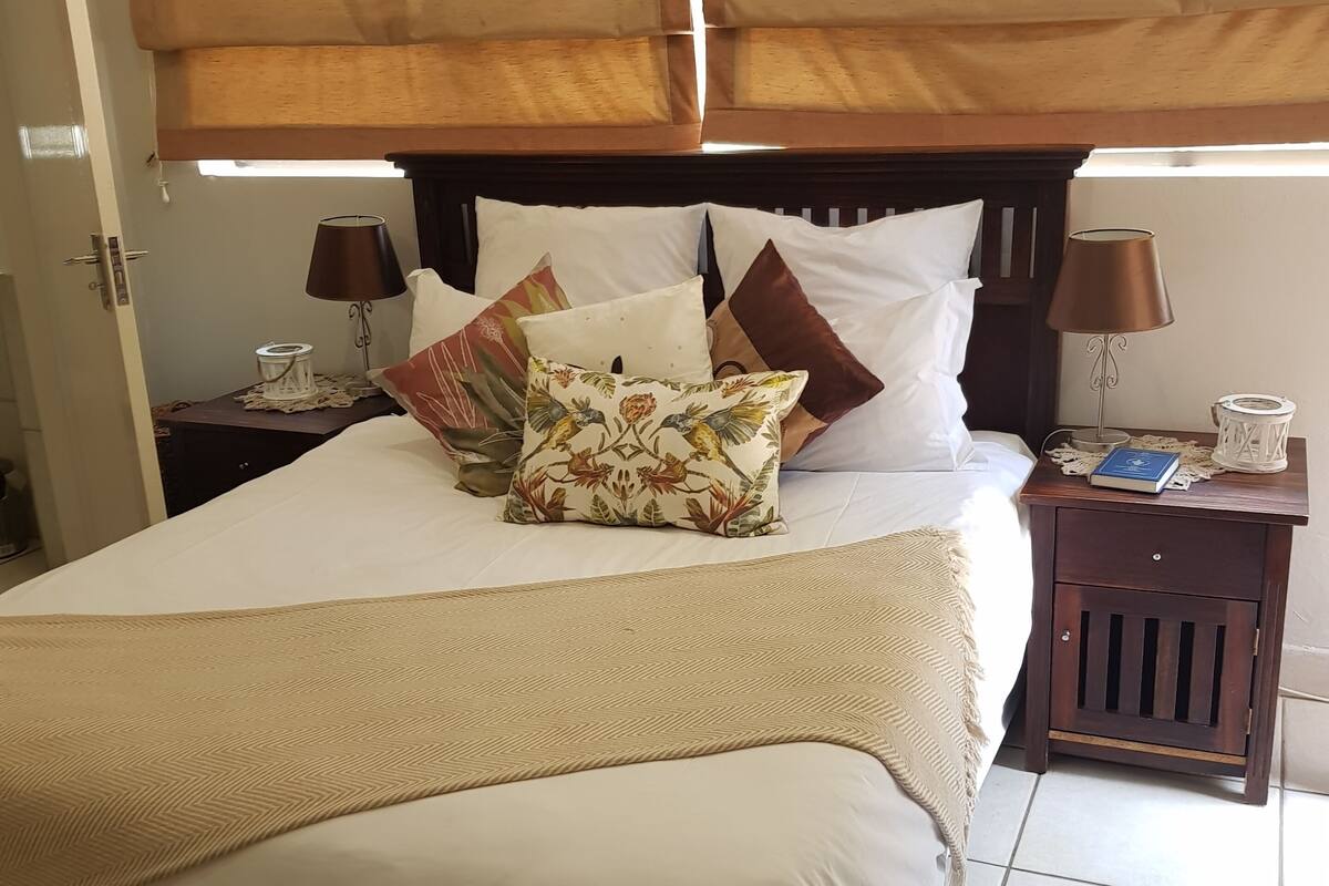 Cama bem arrumada com cabeceira, duas mesas de cabeceira com luminárias, e uma mesa de cabeceira com um livro e um objeto decorativo.