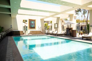 A swimming pool with lounge chairs and a sun umbrella, surrounded by a covered area with a view of trees and a building with patterned windows. - Hotel Hacienda Aguacaliente (Tijuana)