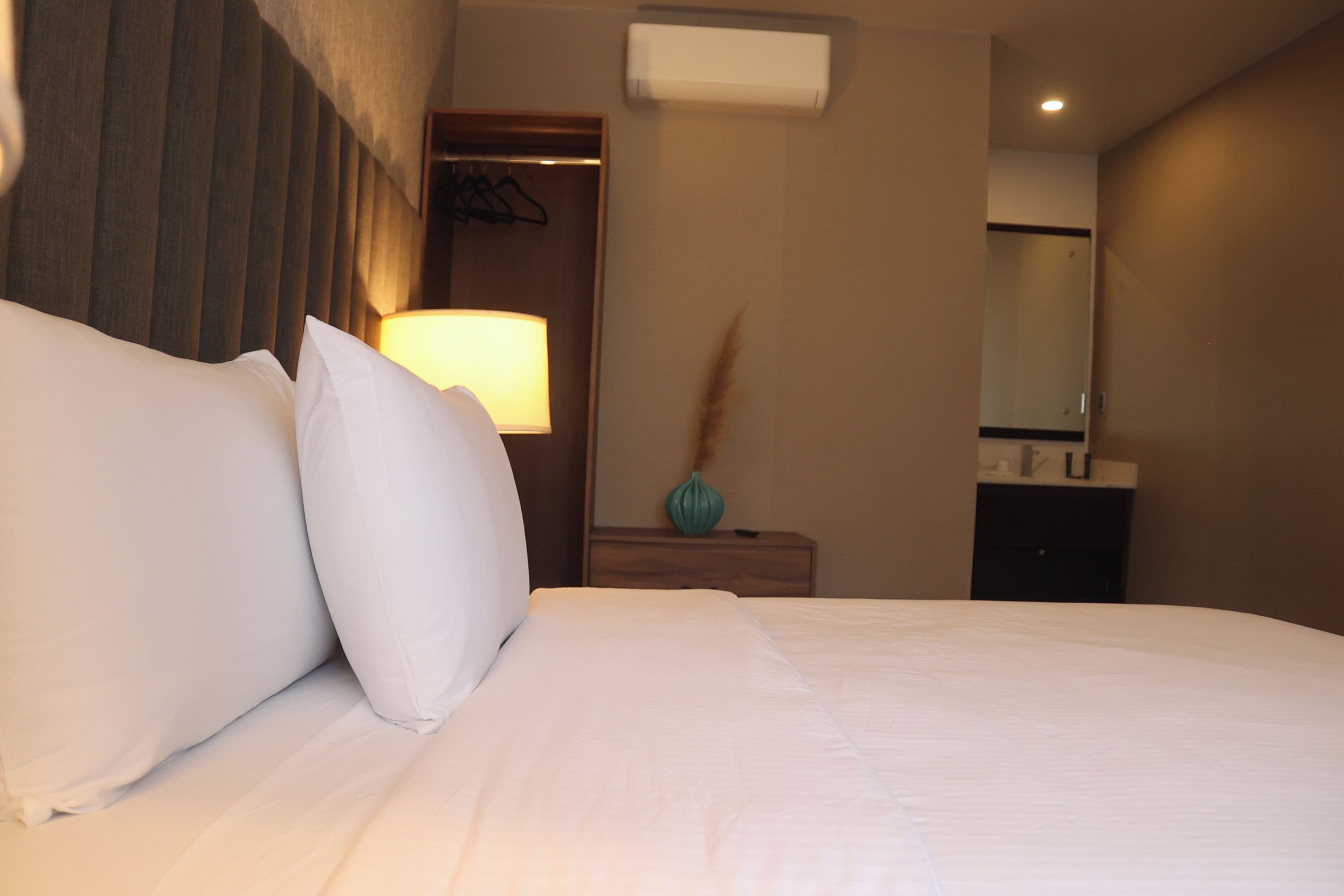 Habitación de hotel con una cama bien hecha, una lámpara de noche, una mesita con un florero y un espejo en la pared.