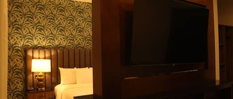 A hotel room with a large flat-screen TV mounted on the wall, a bedside table with a lamp, a bed with white bedding, and a patterned wallpaper on the wall.