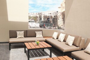 Un área de descanso moderna con un sofá seccional color beige, una mesa de centro de madera con un jarrón de flores y un ventanal con cortinas estampadas.