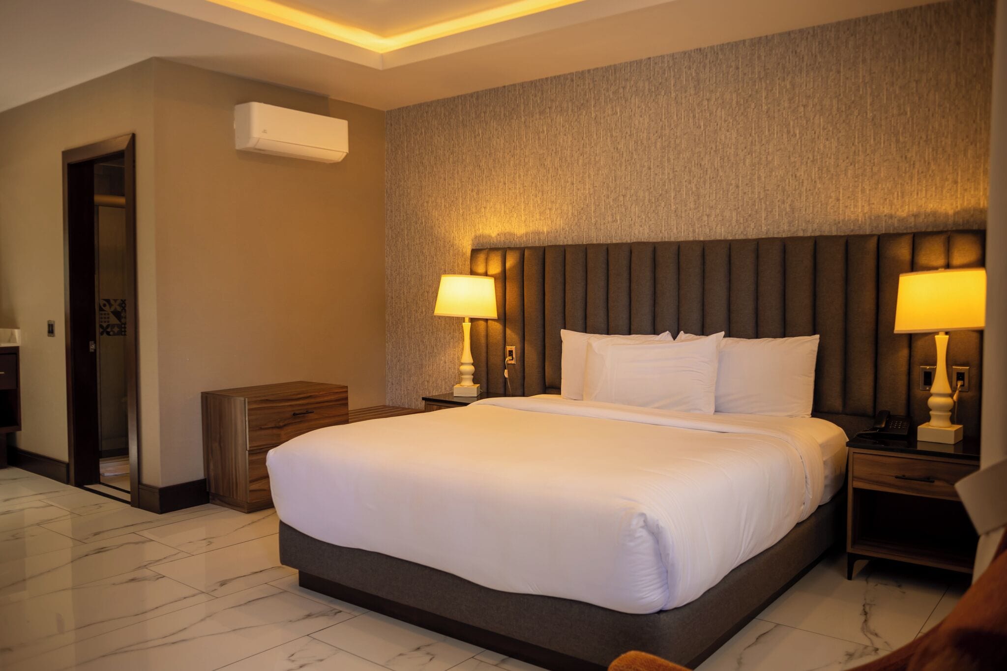A hotel room with a large bed, two bedside tables with lamps, a wooden nightstand, a built-in wardrobe, and an air conditioning unit on the wall.