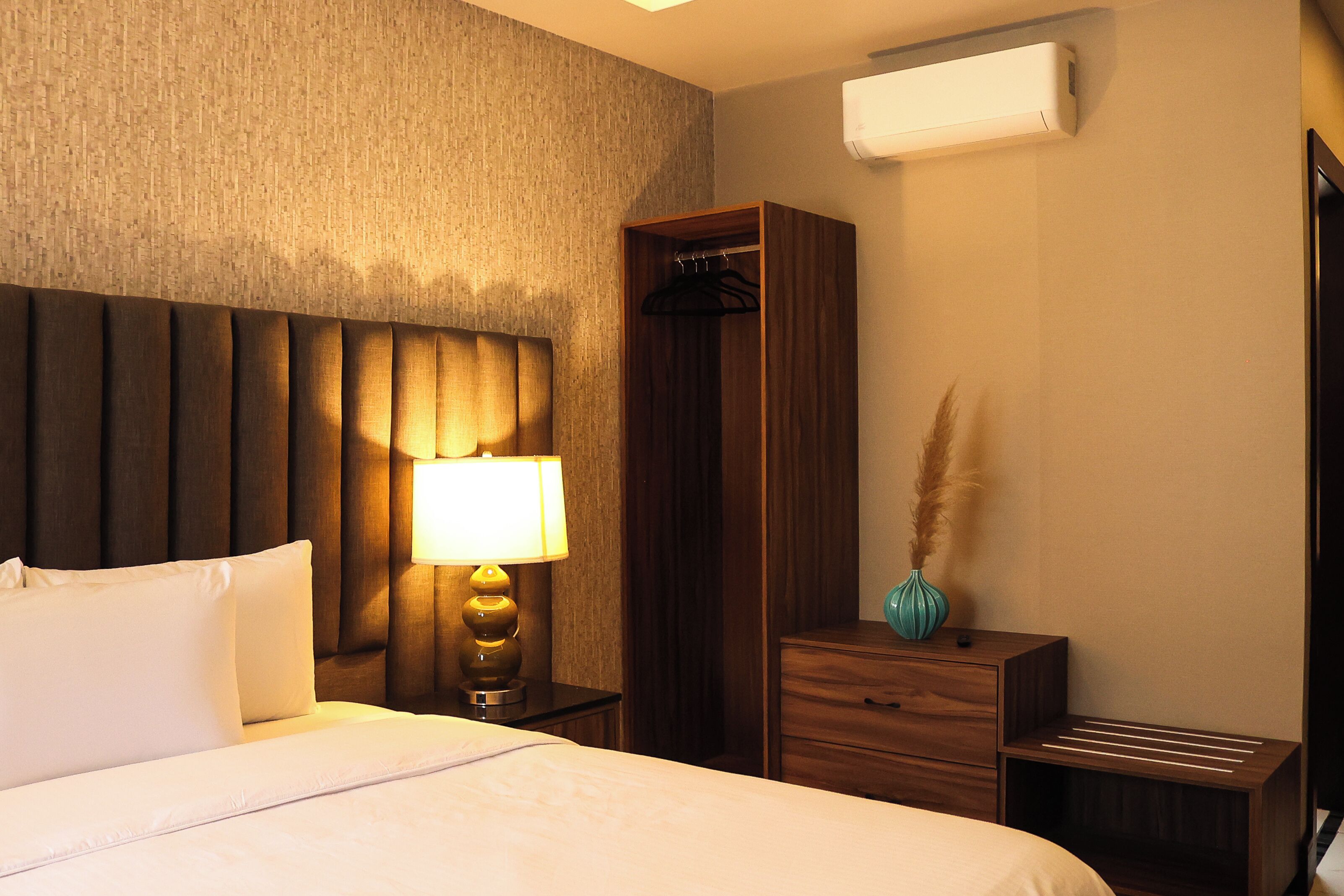 A hotel room with a bed, a nightstand with a lamp, a wardrobe, a small table with a vase, and an air conditioner on the wall.