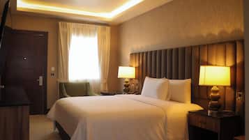 A hotel room with a large bed, two bedside lamps, a wooden headboard, a green chair, and a wooden desk.