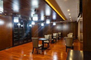 Vestíbulo de hotel con suelo de madera, varias mesas y sillas, e iluminación colgante.