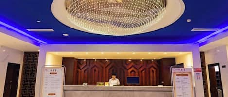 A hotel reception area with a modern design, featuring a large chandelier, a reception desk, and informational stands.