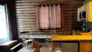 Interior rústico de cabana com mesa de jantar de madeira, duas poltronas de balanço, área de cozinha com micro-ondas e janela com cortinas.