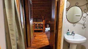 Um corredor de madeira leva a um quarto com uma cama e um pequeno baú de madeira. Uma pia branca com espelho e artigos de higiene pessoal é visível no quarto ao lado.