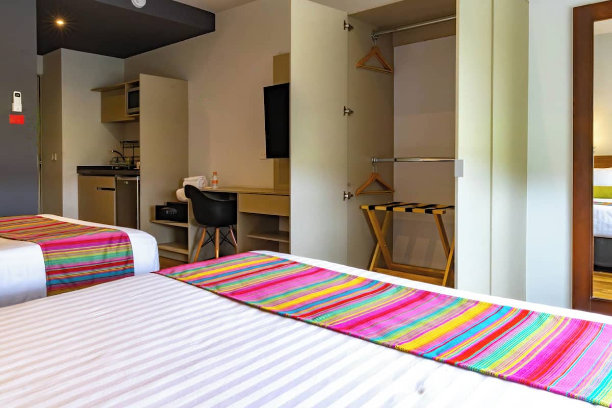 Une chambre d’hôtel avec deux lits, un bureau, une chaise, une armoire et une petite kitchenette.