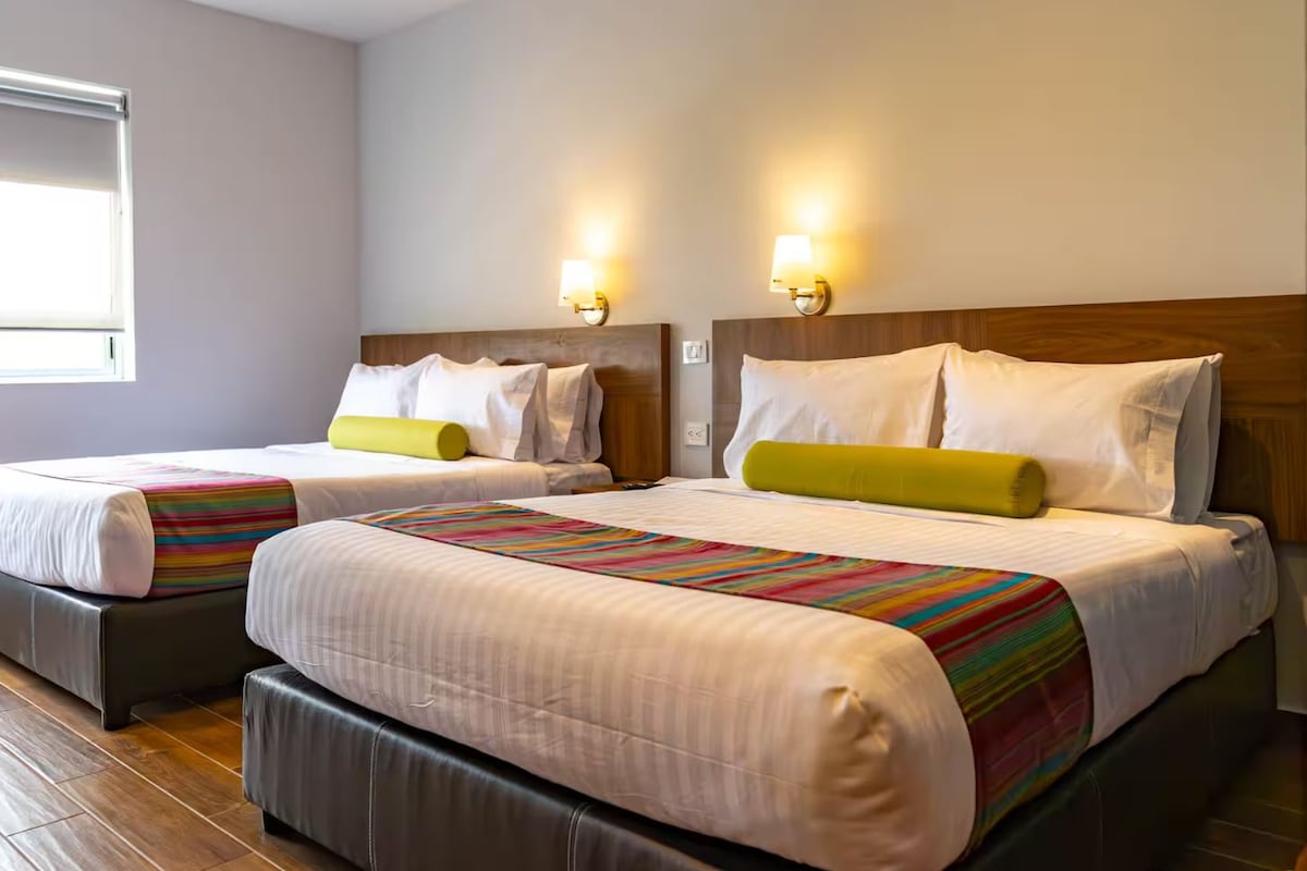 A hotel room with two beds, each with a headboard, a window, and wall-mounted lights.