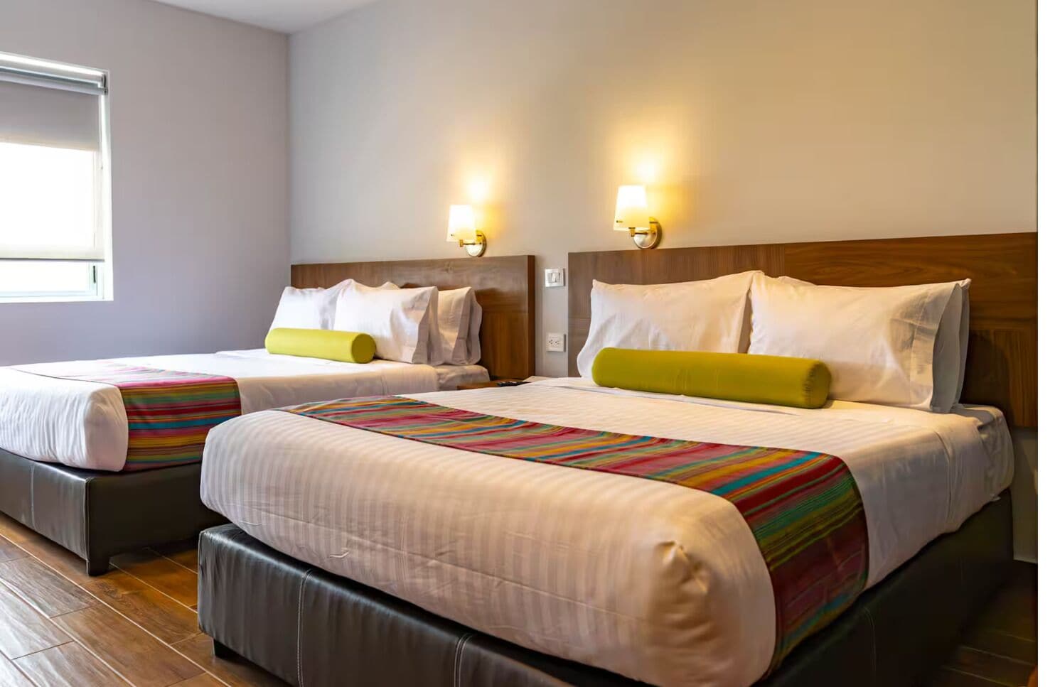 Ein Hotelzimmer mit zwei Betten, jeweils mit Kopfteil, einem Fenster und Wandleuchten.