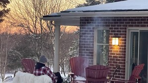 A snow-covered outdoor area with a brick building, a fire pit, and several Adirondack chairs.