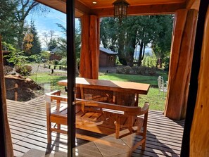 A wooden deck with a table and chairs, overlooking a garden with trees and a building in the background. - cabañasmapulauquen (Villarrica)