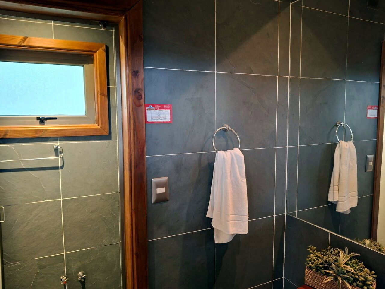 Un baño con paredes de baldosas oscuras, una ventana con marco de madera y dos toalleros con toallas blancas.