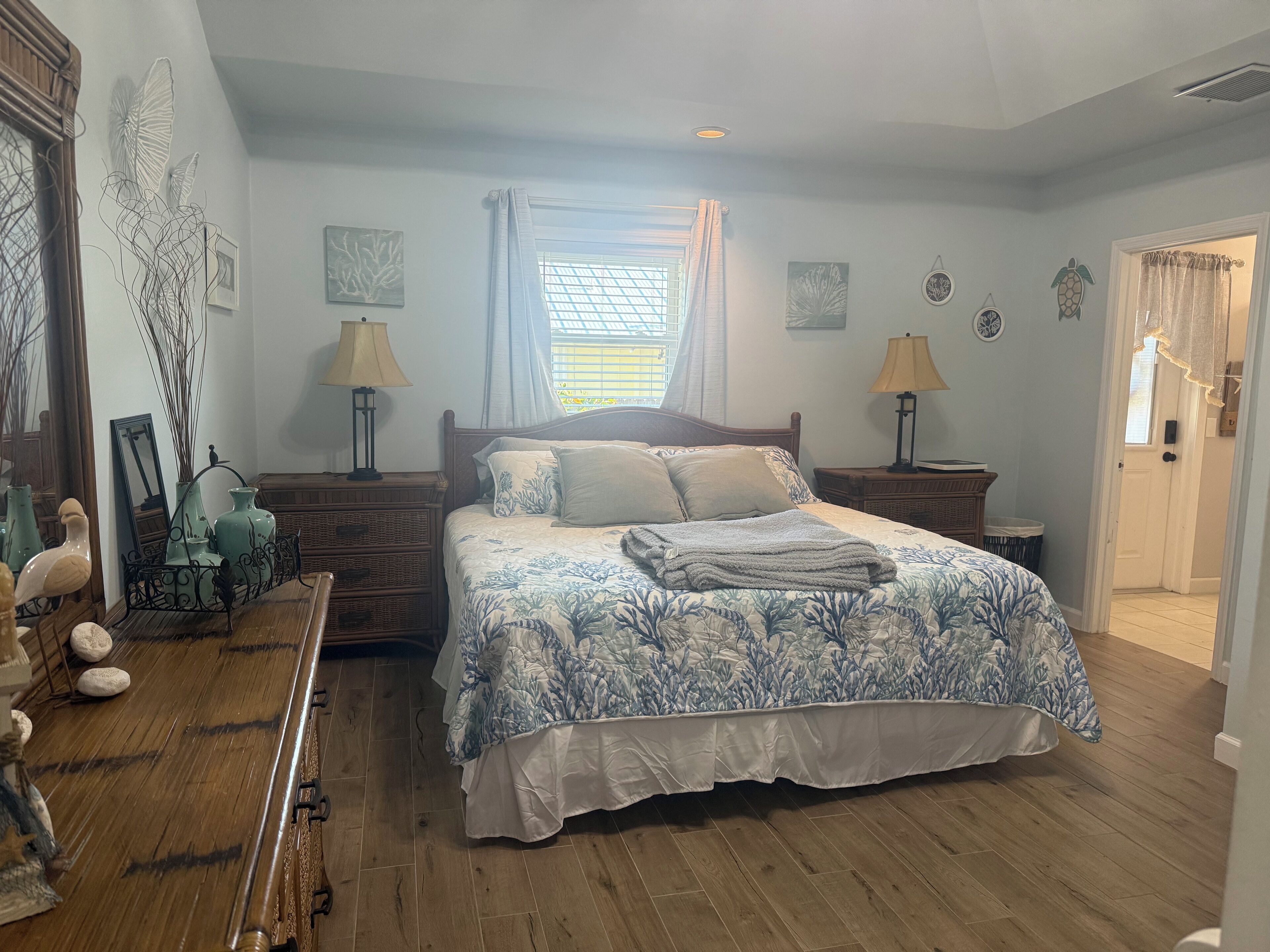 A bedroom with a wooden floor, a bed with a floral bedspread, two bedside tables with lamps, a window with curtains, and wall art.