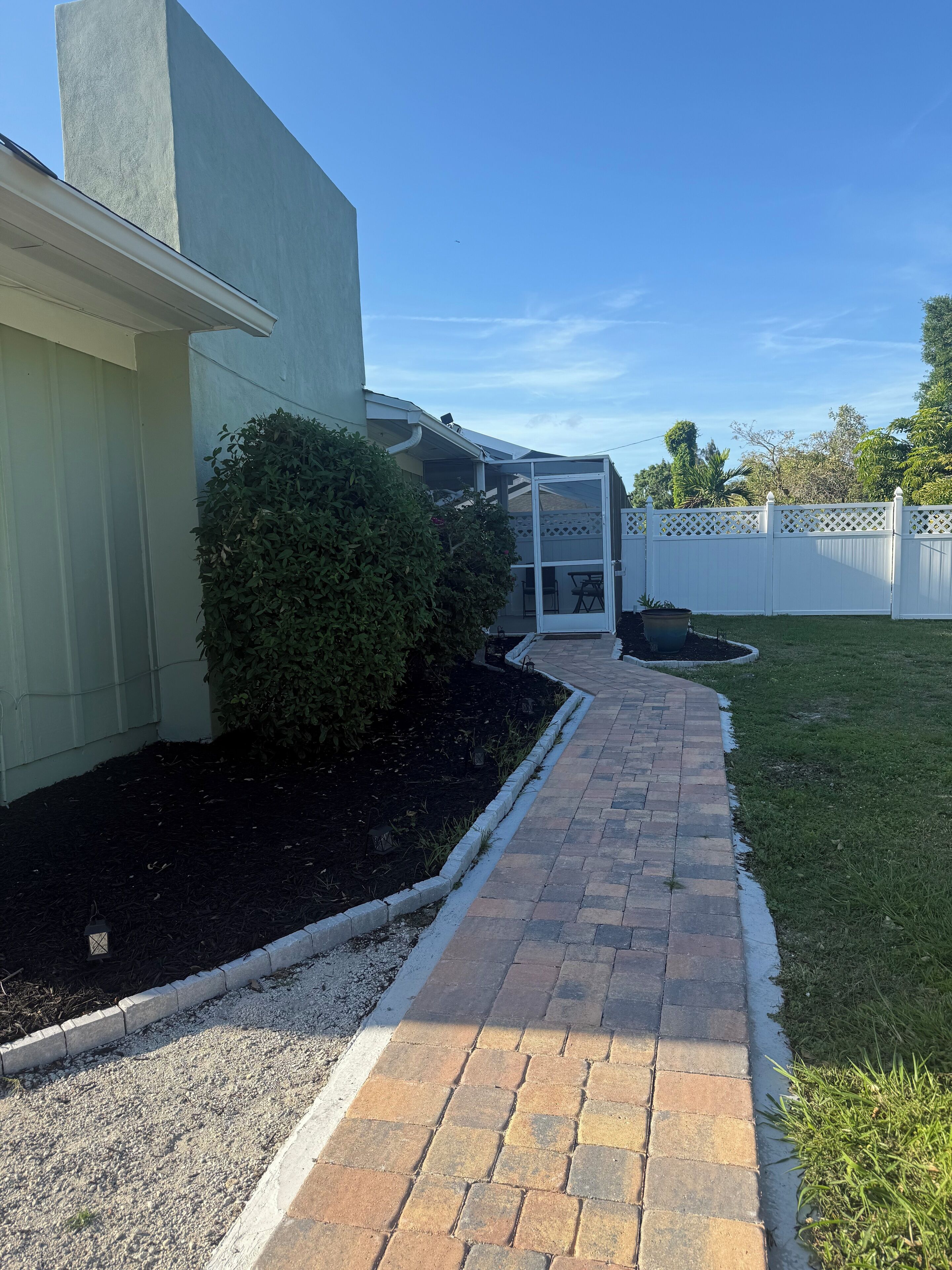 A paved pathway leading to a covered patio area with a white fence and greenery.