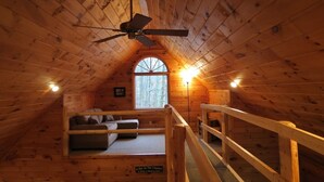 Een gezellig slaapkamer op zolder met houten plafond, een plafondventilator, een groot raam, een sofa en een trap.