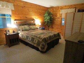 A cozy bedroom with a wooden wall, a bed with a patterned bedspread, a nightstand with a lamp, and a large window with curtains.