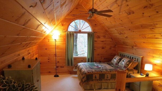 Een gezellig slaapkamer op zolder met een houten schuine dak, een enkel bed met quilt, een nachtkastje met lamp en een groot raam met gordijnen.