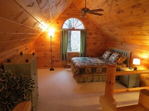 Ein gemütliches Schlafzimmer im Dachgeschoss mit schrägen Holzwänden, einem Bett mit Patchworkdecke, einem Nachttisch mit Lampe und einem großen Fenster mit Vorhängen.