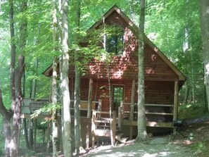 A wooden cabin with a porch, surrounded by tall trees in a forest.