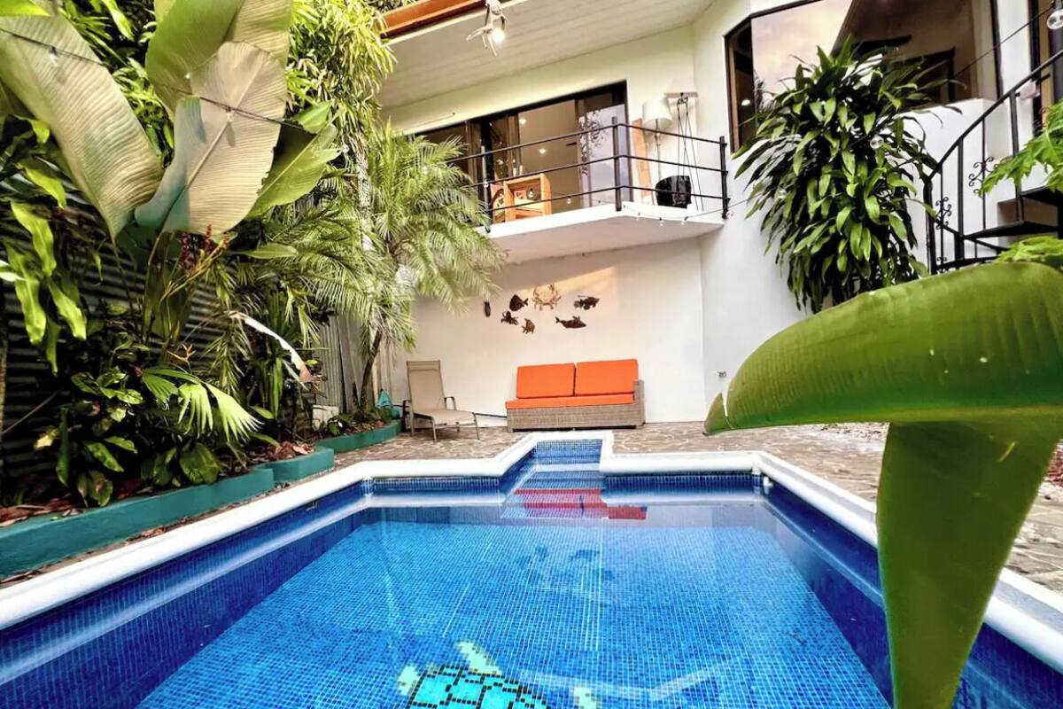 A swimming pool with a tiled edge, surrounded by lush greenery and a balcony with a railing.