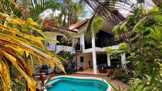 Une piscine entourée d’une végétation tropicale luxuriante, un bâtiment à deux étages doté de balcons et un ciel d’un bleu éclatant.