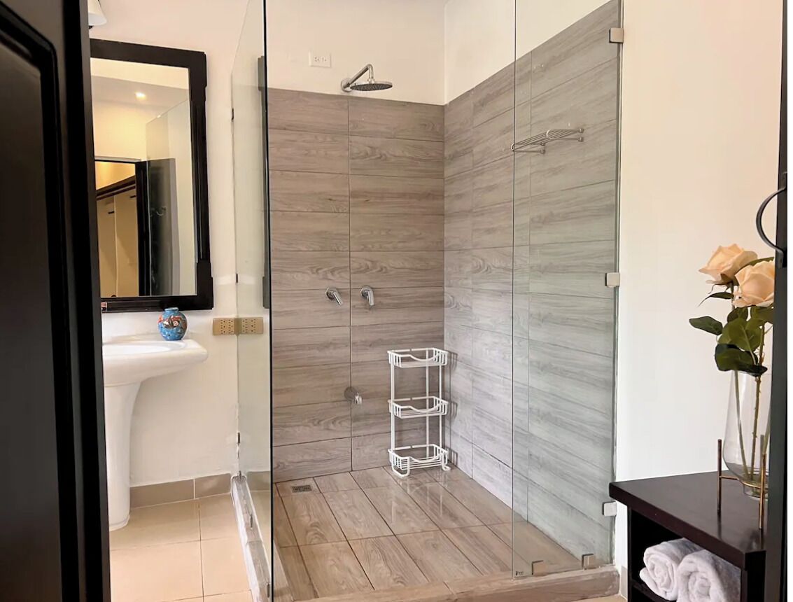A modern bathroom with a glass shower enclosure, a white sink, and a glass vase with flowers on a dark wooden vanity.