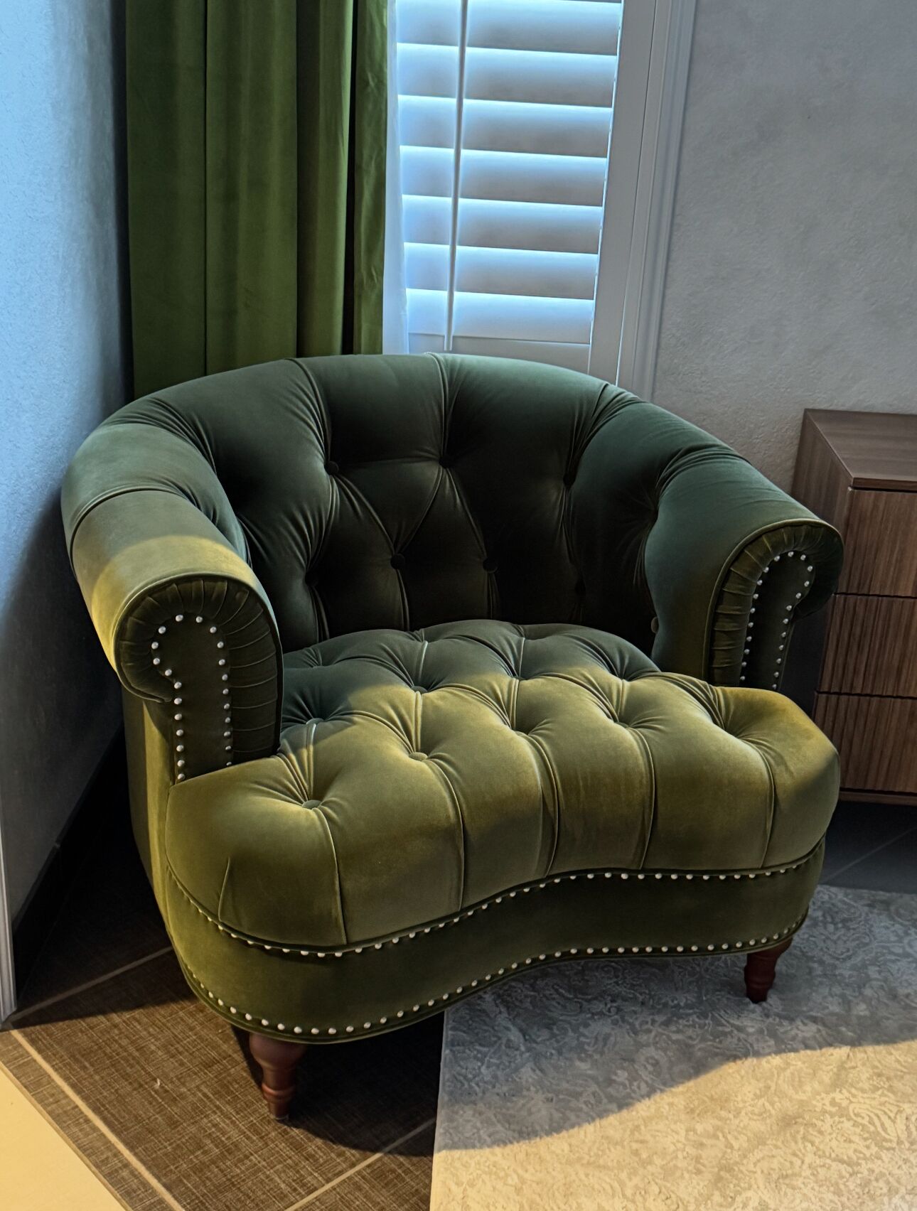 Un fauteuil capitonné doté d’un dossier incurvé et de clous décoratifs, placé dans un coin, à côté d’un meuble en bois et d’une fenêtre avec des stores.