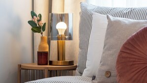 A neatly made bed with striped bedding, a bedside table with a lamp, a vase with flowers, and a large window with curtains.