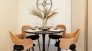 A dining area with a round table, four chairs, a clock on the wall, and a vase with dried plants.