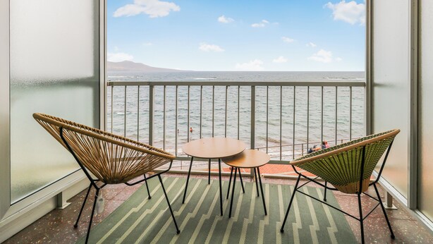 A balcony with two wicker chairs and a small table, overlooking a body of water and a distant mountain.