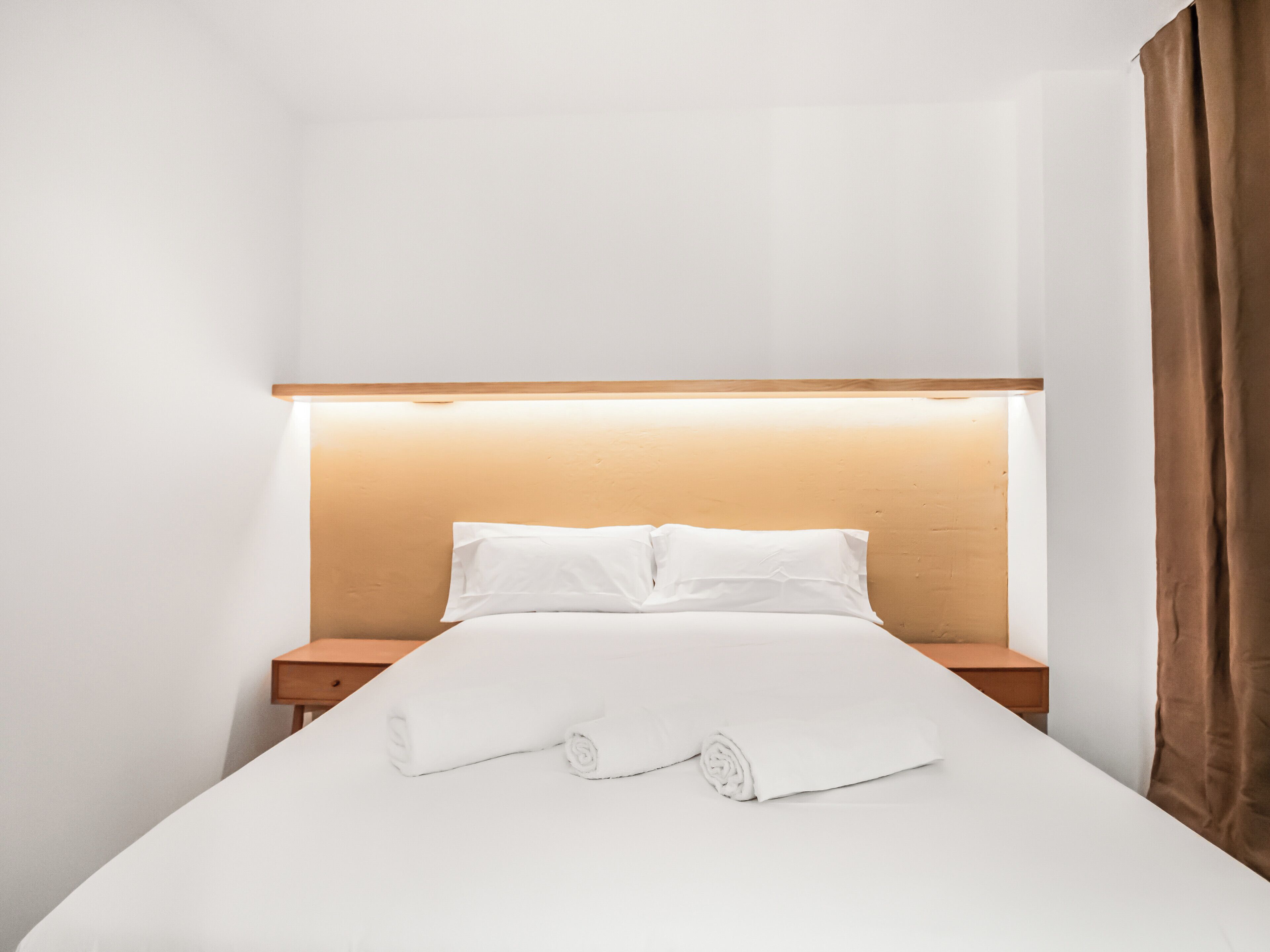 Ein ordentlich bezogenes Bett mit weißen Leinen und einem aufgerollten Handtuch, ein Nachttisch aus Holz und ein Kopfteil mit integrierter Beleuchtung.
