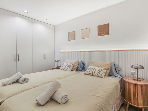Quarto de hotel moderno com duas camas, um guarda-roupa, uma mesa de cabeceira com abajur e obras de arte nas paredes.