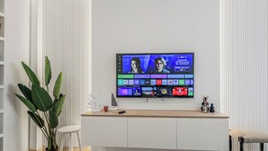 A modern living room with a flat-screen TV mounted on a white wall, a wooden coffee table, a white chair, and a potted plant.