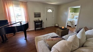 A living room with a grand piano, a cozy sofa, a small table with a candle, and a door leading to a bedroom. - Cozy Cottage in the City (Saint Ann)