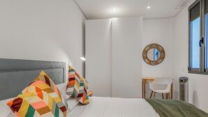 A modern bedroom with a bed, colorful pillows, a desk, a chair, a mirror, and a window.