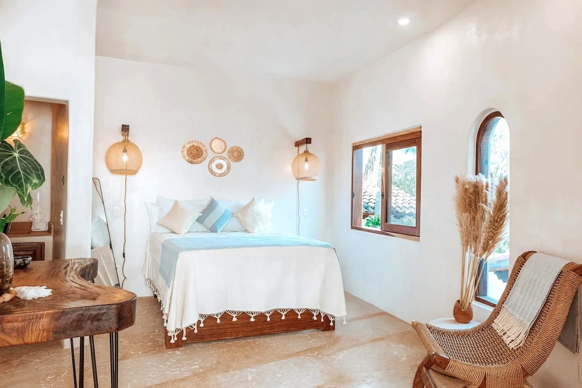 Un dormitorio con cama, lámparas de noche, una ventana, una silla de mimbre y una mesa con un jarrón.