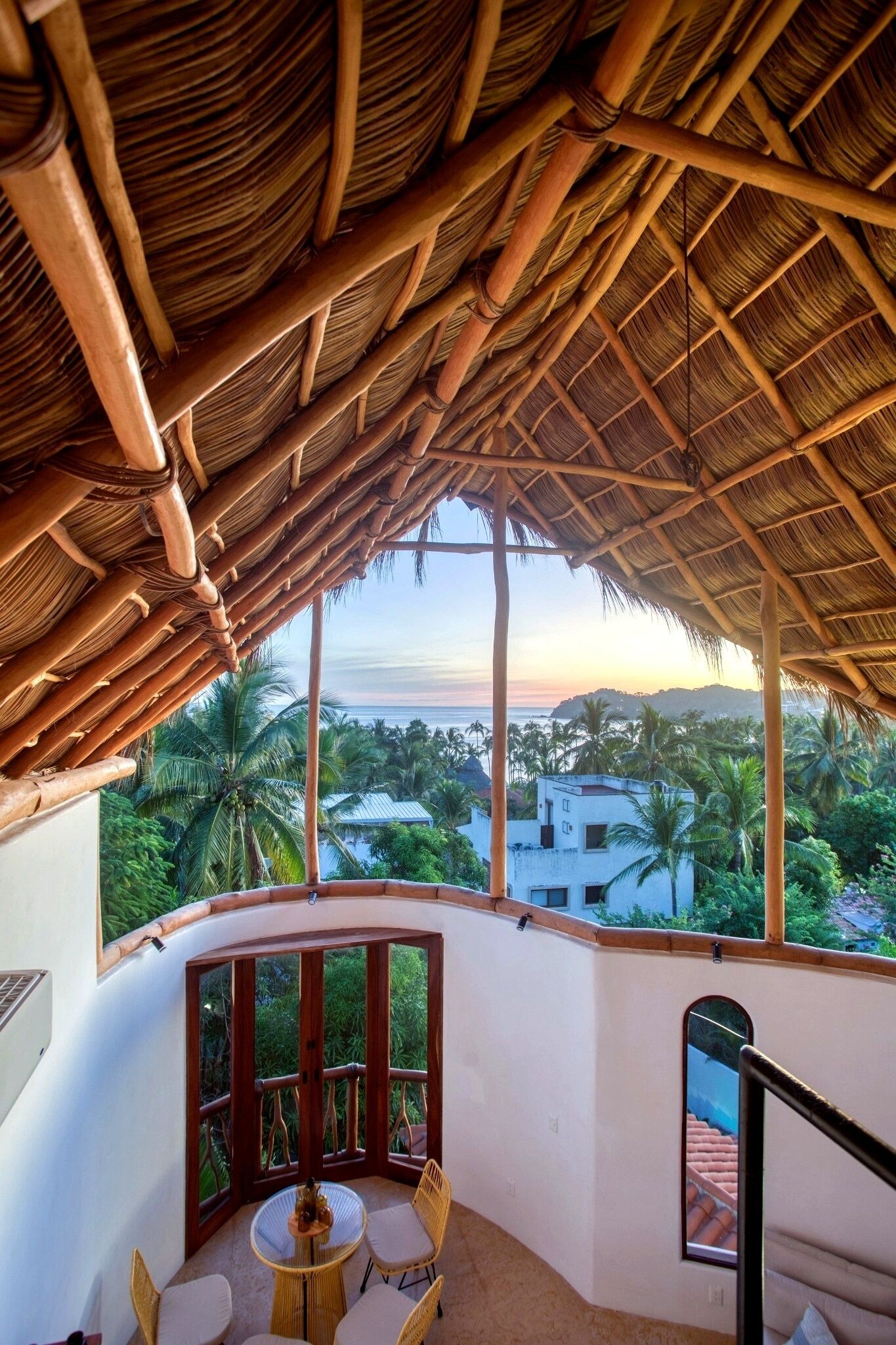 A room with a thatched roof, large windows, and a view of palm trees and buildings.