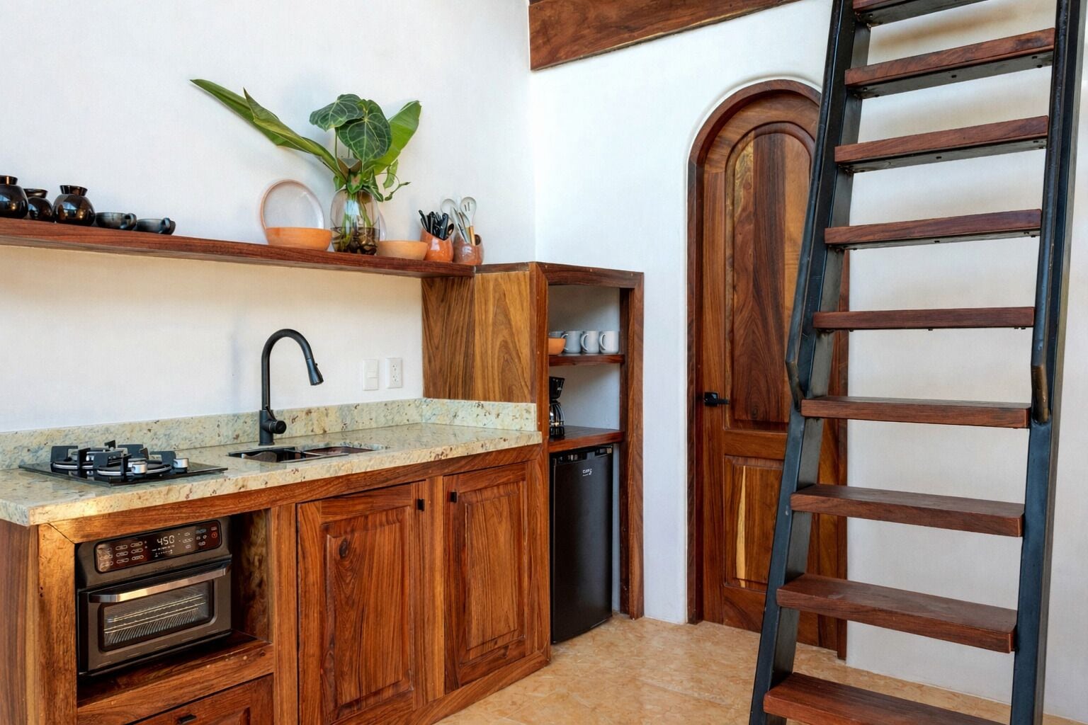 A compact kitchen with wooden cabinets, a built-in oven, a coffee machine, and a sink. There is a plant on the countertop and a ladder against the wall.