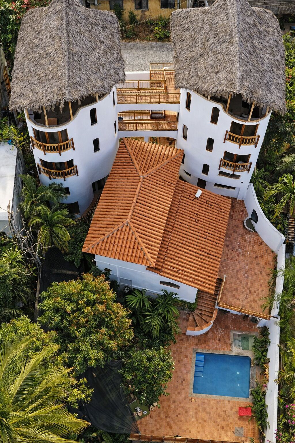 Um edifício com vários andares, telhado de cerâmica, duas estruturas com telhado de palha e uma piscina.