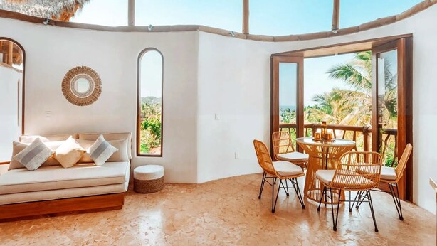 A room with a sofa, a round table with chairs, large windows, and a thatched roof. - Amari Boutique Hotel Sayulita (Sayulita)
