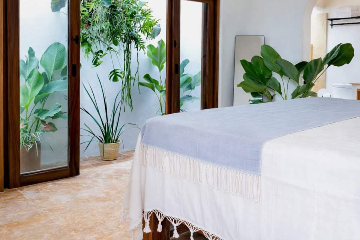 Un dormitorio con una cama, un ventanal corredera que da a un balcón con plantas y un equipo de aire acondicionado en la pared.