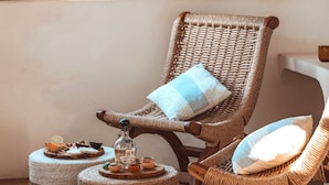 A wicker chair with a blue pillow, a round ottoman with a bottle, and a wooden shelf with bottles. - Amari Boutique Hotel Sayulita (Sayulita)